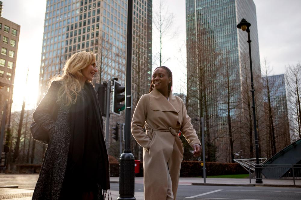 Duas mulheres caminhando e conversando em uma rua urbana, com prédios comerciais altos ao fundo, em um ambiente corporativo moderno