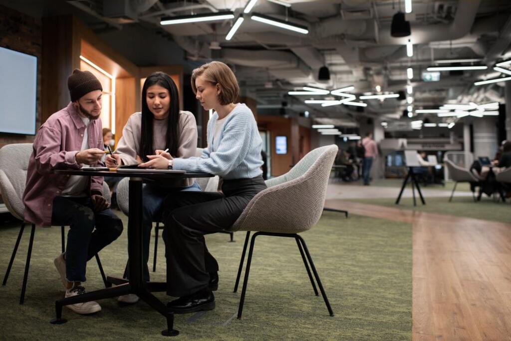 Imagem de três pessoas jovens em um ambiente moderno de coworking, discutindo e olhando para o celular, com iluminação sofisticada e atmosfera colaborativa.