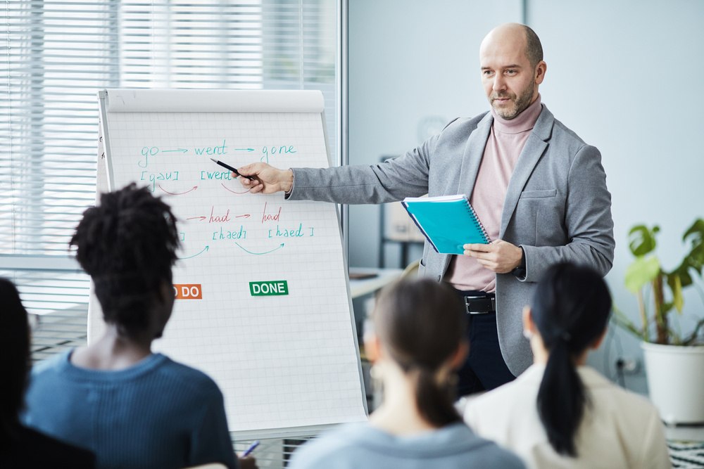 Homem apresentando informações de inglês para negócios enquanto aponta para anotações sobre verbos no flip chart.
